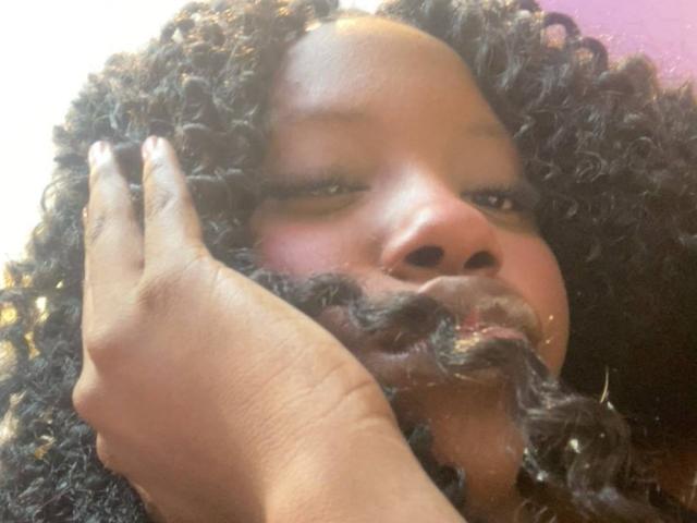 CatalinaSanA, avec ses cheveux noirs et bouclés, porte sa main devant son visage et a une énorme moustache brun-grisâtre qui lui recouvre la bouche. Sa peau est foncée et elle a des ongles blancs..