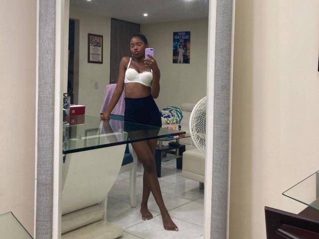 CatalinaSanA with dark skin, small white bra, and black mini-skirt, standing barefoot in front of mirror, holding phone, showing her tits and ass. Room has glass table.