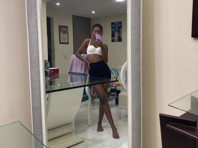 CatalinaSanA, dark-skinned, standing in front of mirror, wearing white bra and black skirt, holding phone, purple towel on chair, white fan, glass table, wooden dresser in background.