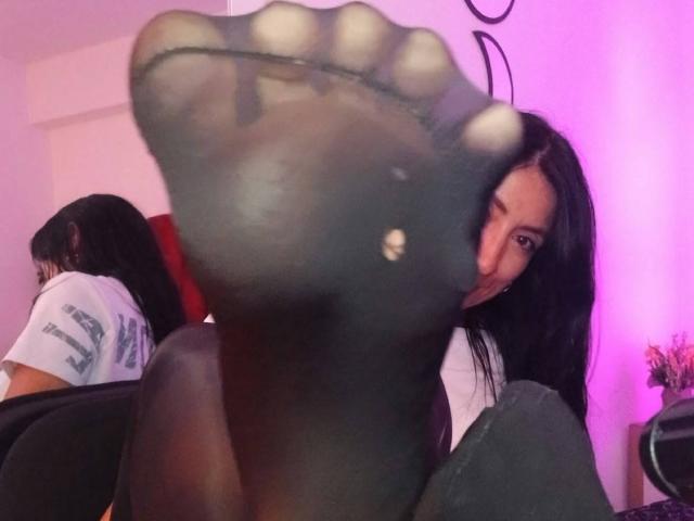 ZarayFeet's foot, showing a small hole in her sheer black sock, with her black hair and a slight smile in the background, her ass and tits partly visible.