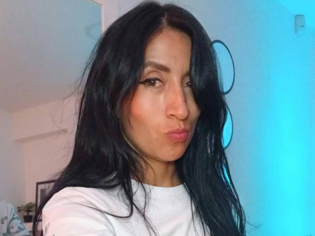 ZarayFeet has long black hair, pouty pink lips, and is wearing a white shirt. She's making a pouty face, with a slight smile, in a blue-lit room.