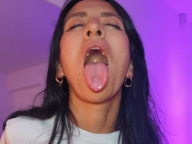 ZarayFeet's face with eyes closed, mouth open, tongue out, showing the roof of her mouth and slightly wet tongue. She has long black hair, hoop earrings, and a white shirt.