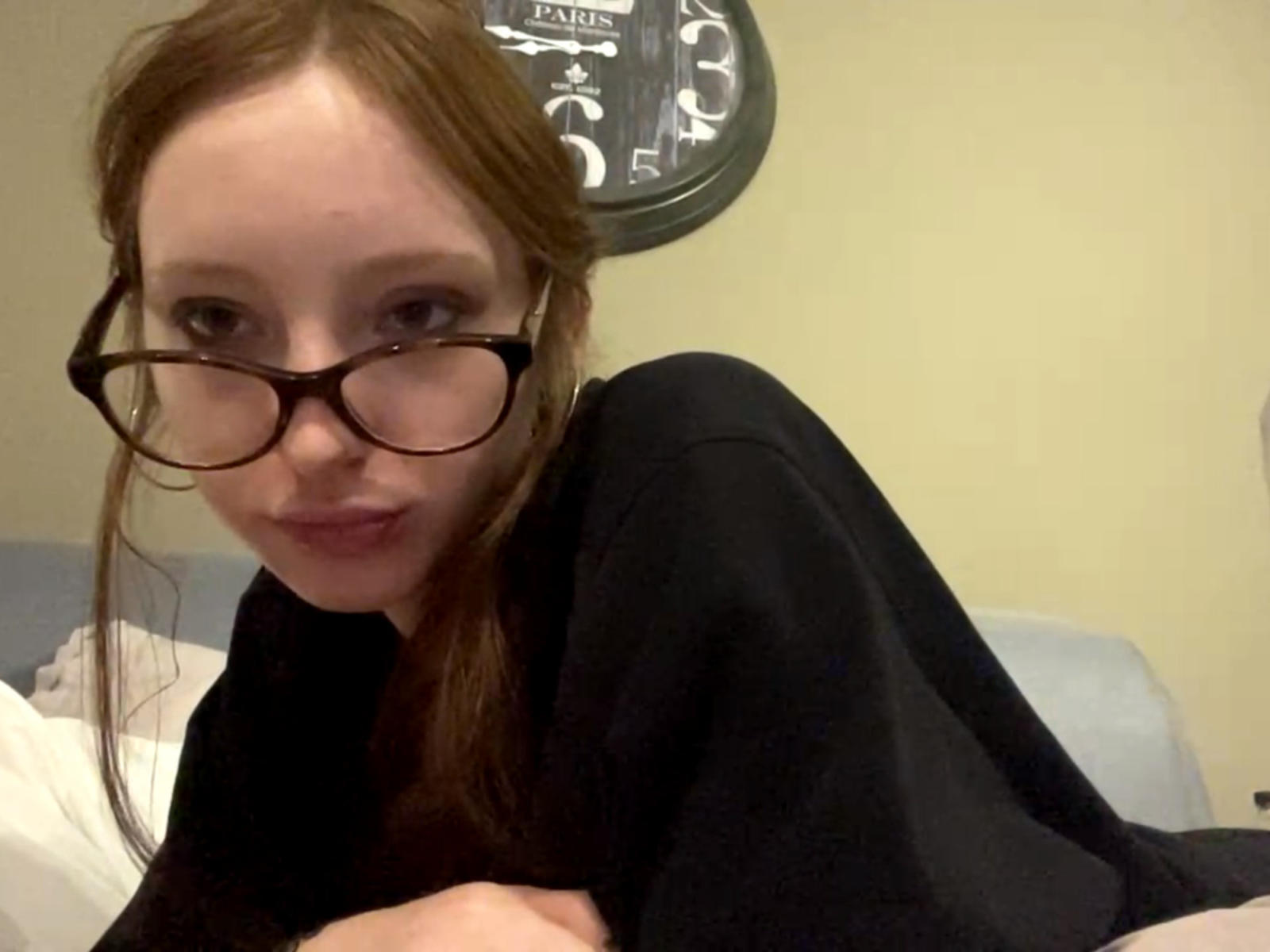 MaeganCepero with glasses, red hair, black top, and a slight smile, lying on a bed, looking at the camera, arms crossed. Her ass is partially visible.