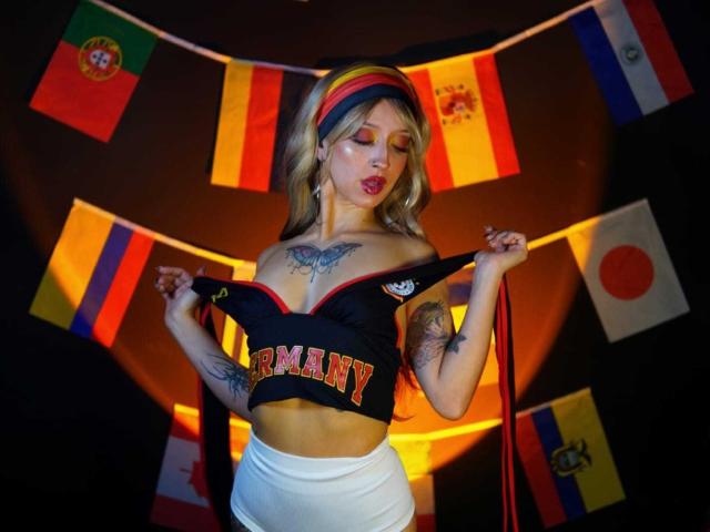 AnaRossii with blonde hair, wearing a black "GERMANY" crop top, white pants, and rainbow headband, showing tattoos, holding top, red lips, looking down, colorful flags in background.