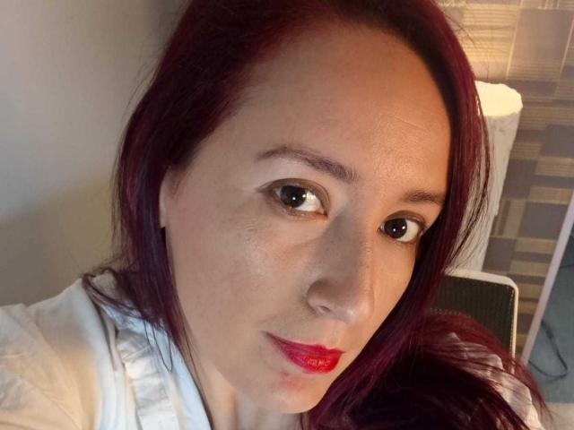 A SweettEve tem cabelo ruivo, pele clara e batom vermelho. Seus olhos castanhos e a maquiagem discreta são visíveis. Ela está vestindo uma camisa branca e olhando diretamente para a câmera..