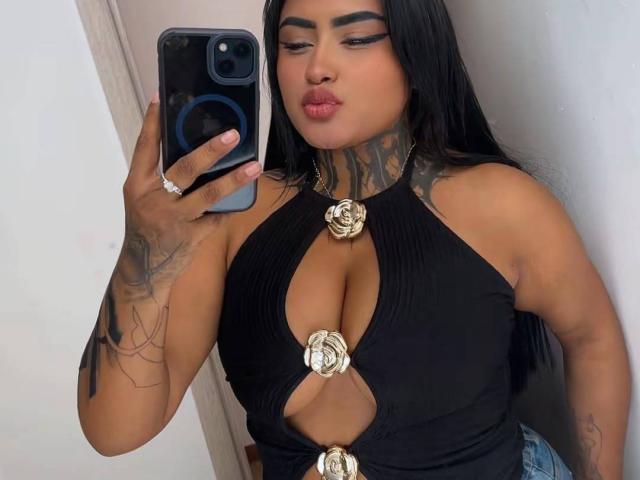 LairaHearvy taking a mirror selfie, showing off her black halter top with gold rose buttons, tattoos on her neck and arm, and pouting her plump lips. Her hand has a ring.