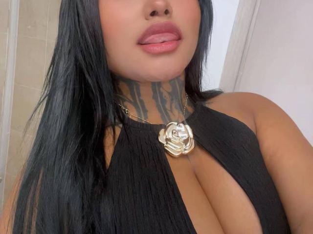 LairaHearvy with black hair, tongue out, wearing a black halter dress with a rose necklace, showing large tits, and a tattooed neck. Her lips are pink and glossy.