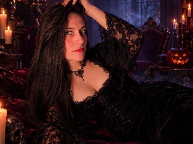 SaraGraham with long black hair, wearing a black lace dress, exposing her tits, lying on a red velvet couch, hand in hair, surrounded by candles, in a dark room.