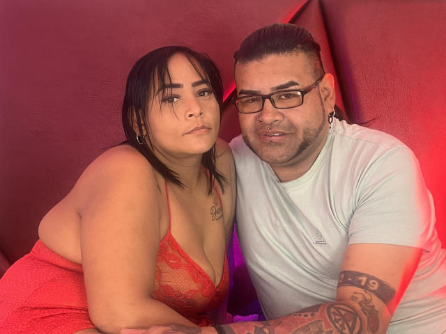 RedFireMoon with big tits in red lace dress, and RedFireMoon with glasses, tattoos, and white tee, leaning together, red and purple lighting, slight smirk, black hair, and beard.