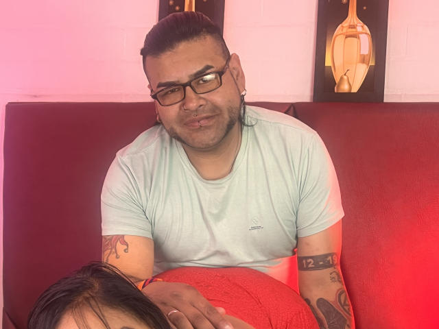 RedFireMoon with tattoos, light green t-shirt, and glasses, pouting, sits on red couch, hand on another person's red-covered ass. Red lighting. Short, dark, slicked-back hair.
