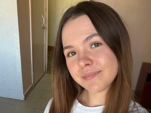 JeseniaPiontek has brown eyes, straight brown hair, and a slight smile. She's wearing a white shirt, with a small mole on her right cheek and no makeup.