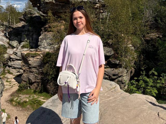 JeseniaPiontek with long brown hair, pink t-shirt, denim shorts, white handbag with purple straps, standing on rocky terrain, wearing black sunglasses on head, smiling, sunny day.