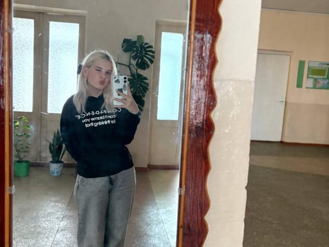 FallenAngelia with blonde hair, black sweater, and gray pants, taking a mirror selfie in a room, holding a phone, showing her ass in the reflection.