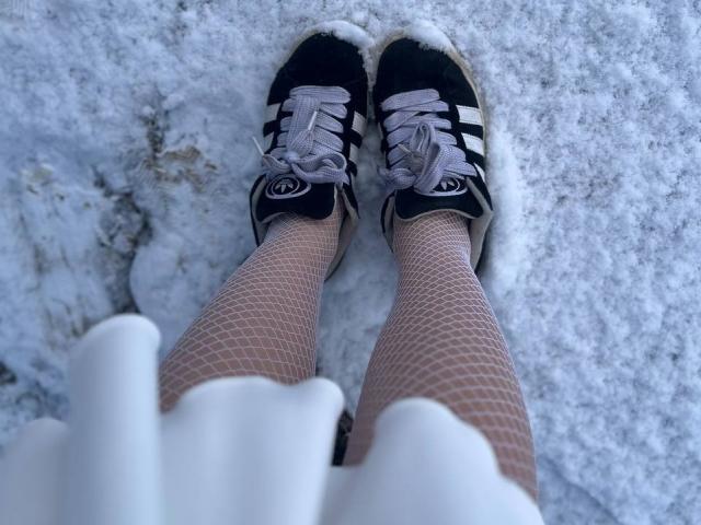 FallenAngelia's legs in fishnet stockings, wearing black and white Adidas sneakers with white laces, standing on a snowy, textured ground. Her white skirt is partially visible.