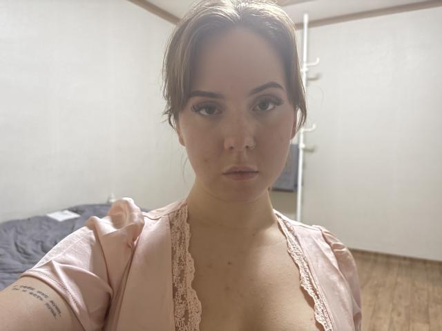 NonaClaudio with light skin, brown hair in a loose updo, wearing a pink lace-trimmed top, showing cleavage, looking at the camera with a neutral expression, in a room.