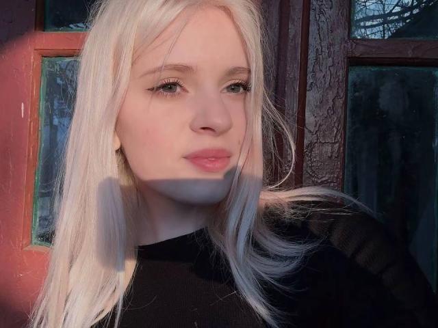 FallenAngelia with platinum blonde hair, light skin, and pink lips, wearing a black top, looking slightly to the side with a soft, serious expression. Sunlight casts a shadow on her face.