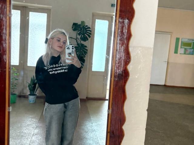 FallenAngelia with blonde hair, black sweater, and grey pants, standing in front of a mirror, taking a selfie with a white phone, in a room with a plant.