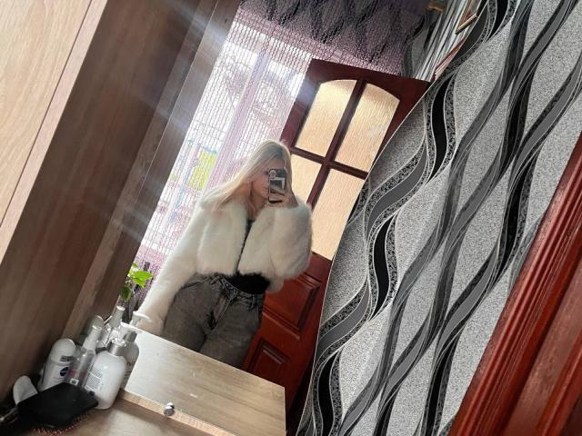 FallenAngelia with blonde hair, wearing a fluffy white fur jacket and high-waisted gray pants, taking a mirror selfie, showing her tits and smiling, in a wooden room.