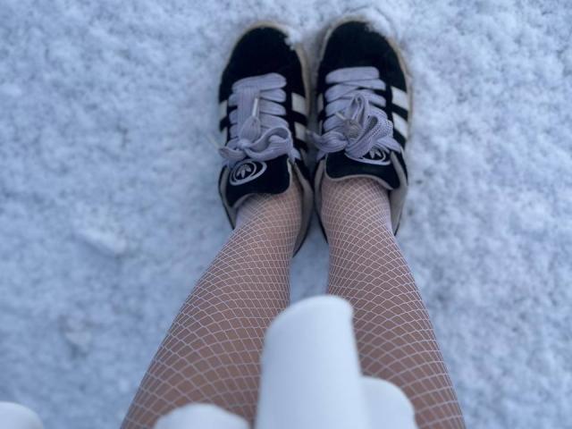 FallenAngelia's legs in fishnet stockings, wearing black Adidas sneakers with white laces, standing on a white, textured, snow-like surface. FallenAngelia's feet are pointed slightly inward.
