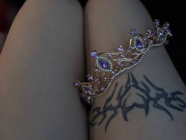 FallenAngelia's pale thighs with a tattoo of black, spiked branches, wearing a gold crown with purple gems, positioned on her upper thigh.