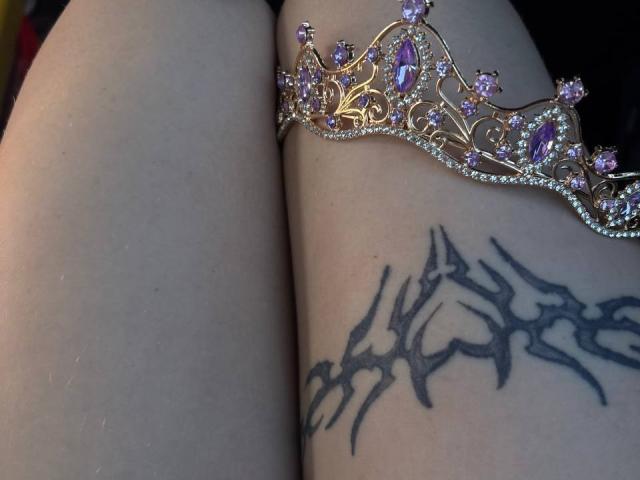 FallenAngelia's thigh with a black tribal tattoo, next to a gold crown adorned with purple and pink gemstones, resting on her pale skin.