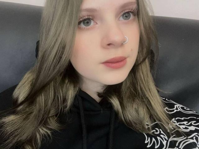  FallenAngelia with pale skin, green eyes, and brown wavy hair, wearing a black hoodie with white patterns, has a small nose ring and pink lips.