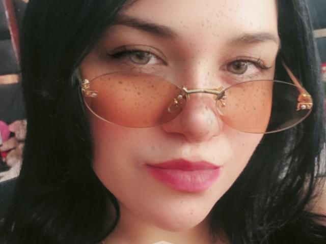 Viviana Thomson, avec sa peau constellée de taches de rousseur, ses cheveux noirs et ses lèvres roses, portant des lunettes aviateur à monture dorée, a un visage mignon et confiant. Son petit nez et ses yeux verts sont visibles..