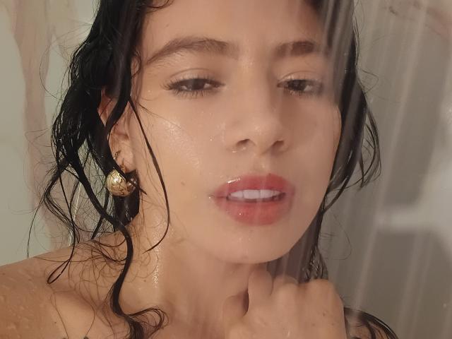 Lindas feuchtes Gesicht, schwarzes, lockiges Haar und glänzende, rosafarbene Lippen mit Wassertropfen, während sie einen Duschvorhang hält. Sie trägt goldene Ohrringe und blickt leicht nach unten..