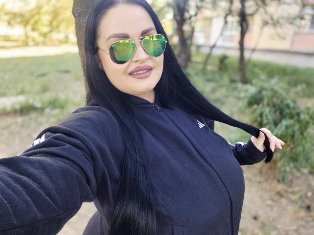 GeorgiaLerox, con lunghi capelli neri, occhiali da sole verdi riflettenti e una giacca nera Adidas con zip, tiene i capelli, sorride, è all'aperto, c'è luce solare, ha gli occhi verdi, labbra piene e una figura leggermente formosa e attraente..