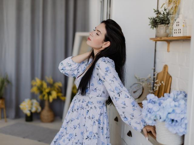 JasmineKouch with long black hair, white dress with blue floral patterns, leaning against white cabinet, hand in hair, eyes closed, standing next to blue hydrangeas, soft smile.