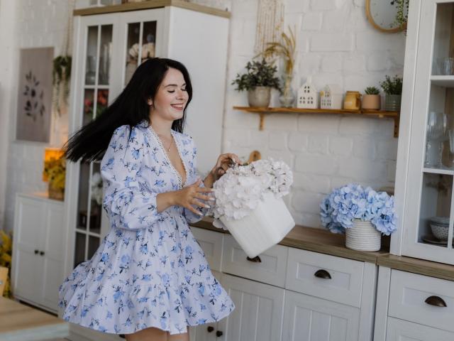 JasmineKouch with black hair, blue floral dress, smiling, holding white flower bouquet, white brick wall, wooden shelves, white cabinets, blue hydrangea, wooden countertop, cozy kitchen.