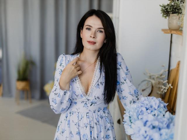 JasmineKouch with long black hair, blue floral dress, lace trim, holding blue flowers, standing near shelf with potted plants, slightly smiling, right hand on chest.