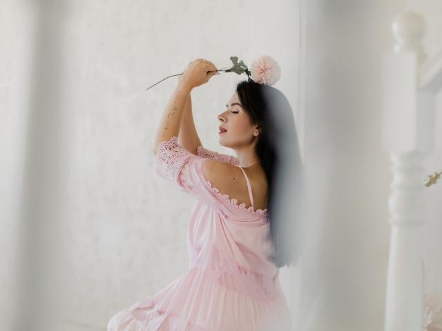 JasmineKouch with dark hair, pink off-shoulder dress, holding pink flower, arms raised, eyes closed, leaning against white wall, soft light, delicate, sexy, pretty, subtle tattoos.