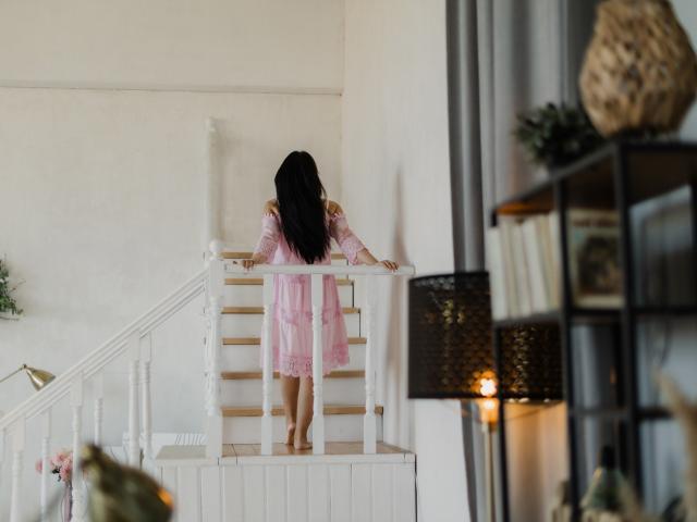 JasmineKouch with long black hair, pink lace dress, and bare feet, climbing white stairs, back to camera, holding railing, soft light, cozy room, wicker lamp on right.