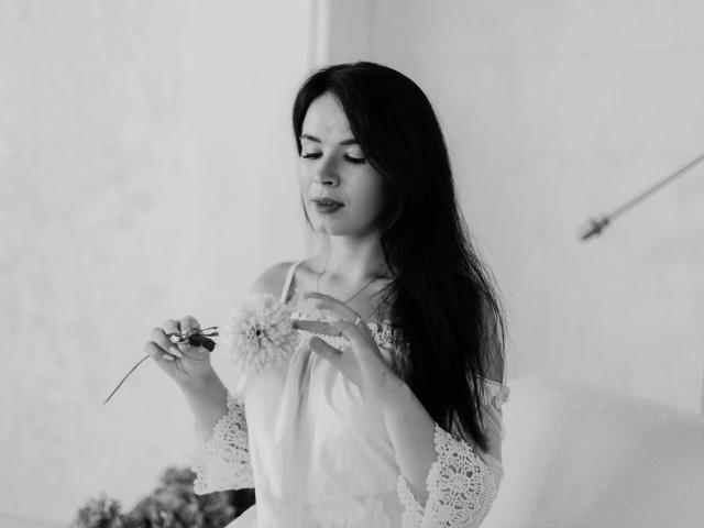 JasmineKouch with long dark hair, holding a daisy, wearing a lace off-shoulder top, looking down, soft light, black-and-white, gentle expression, slight smile, delicate hands.