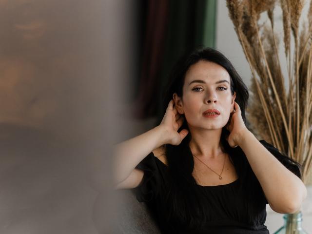 JasmineKouch with black hair, hands in her hair, wearing a black dress, gold necklace, and red lipstick, looking at the camera, puffy ass slightly visible, soft light.
