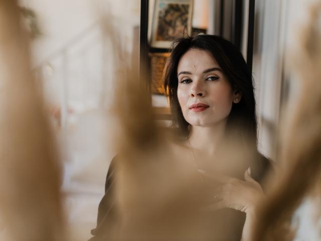 JasmineKouch with dark hair, red lipstick, and subtle makeup, standing in a blurred, warm-toned room, hand near chest, looking slightly to the side, soft and confident expression.