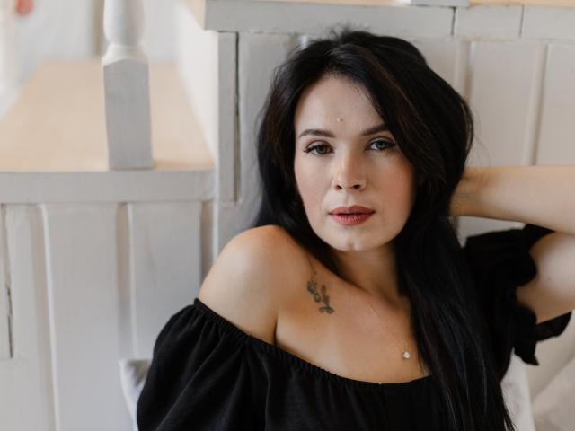 JasmineKouch with long black hair, off-shoulder black top, small tattoo on right shoulder, hand in hair, looking at camera, subtle makeup, white background, soft lighting, relaxed expression.