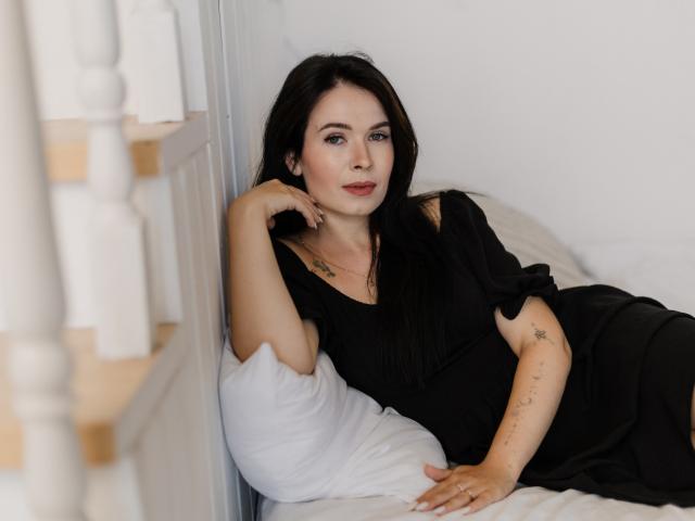 JasmineKouch with black hair, black dress, and tattoos, lying on white bed, hand on pillow, looking at camera, subtle smile, bedroom, soft light, relaxed, sexy.