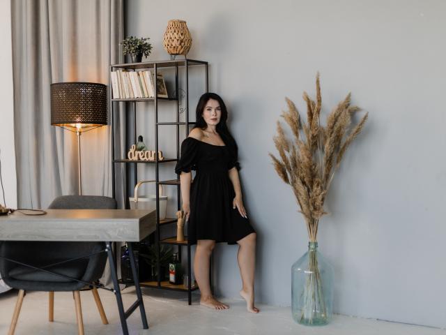 JasmineKouch stands barefoot, wearing a black dress, with her tits slightly visible, next to a puffy reed vase and a metal shelf. Her ass is partly visible.