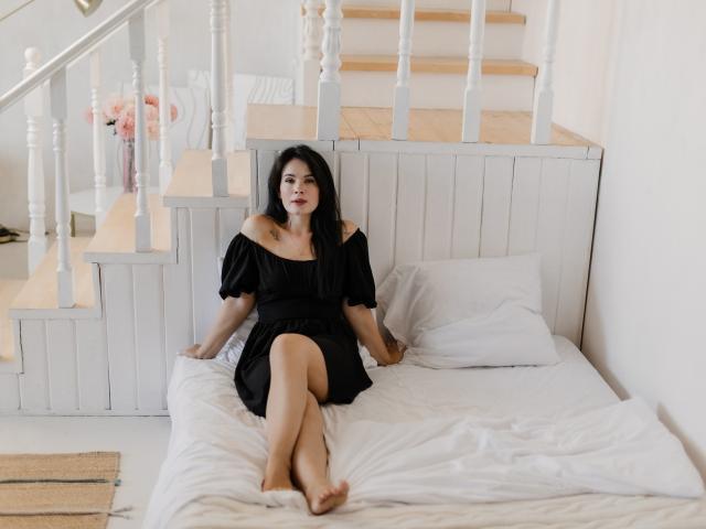 JasmineKouch with long black hair, off-shoulder black dress, and bare feet, sitting on white bed, legs crossed, stairs behind, white walls, light wood, and pink flowers.