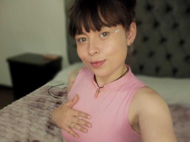 Iniridax with short brown hair, wearing a pink sleeveless top, has small earrings and a pink necklace, touching her tits with one hand, smiling slightly in a bedroom.