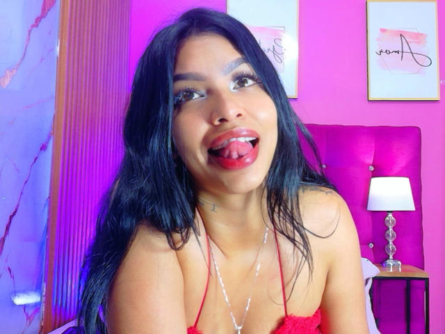 IndianMillion with black hair, sticking out tongue, red lipstick, and cum on chest, wearing red halter top, in pink room, light on side table, headboard in background.