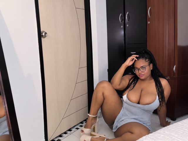 Eilin, avec de grandes seins et des tresses foncées, assise par terre en robe grise, une main dans les cheveux, portant des talons, des lunettes, et regardant l'appareil photo. Chambre avec des portes en bois et un miroir..