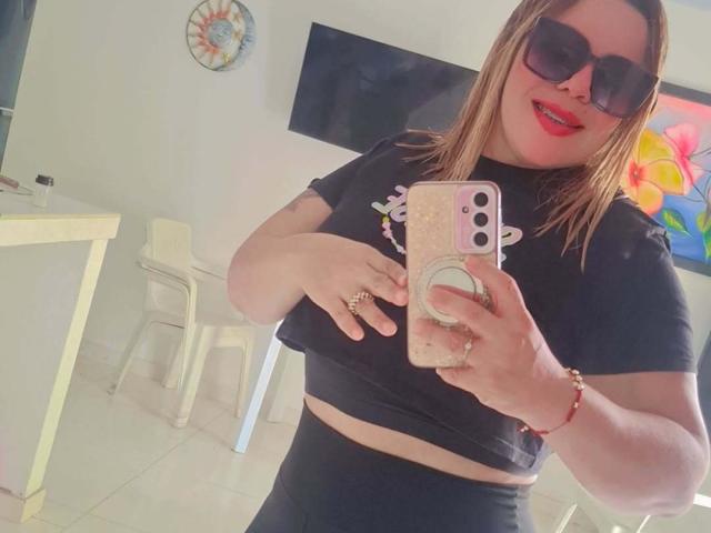 Sharicke69 mit Sonnenbrille, schwarzem Crop-Top und schwarzer Hose, hält ein rosa Handy mit einer Hülle mit einem Ring-Pop, wobei ihre Brüste und roter Lippenstift zu sehen sind. Weißer Raum, weißer Stuhl..