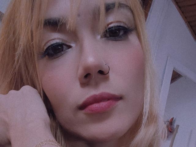 Veroo69 ha i capelli biondi, un piercing al naso e eyeliner, con labbra rosa, guarda verso la fotocamera, con la mano vicino al viso, in un selfie leggermente sfocato..