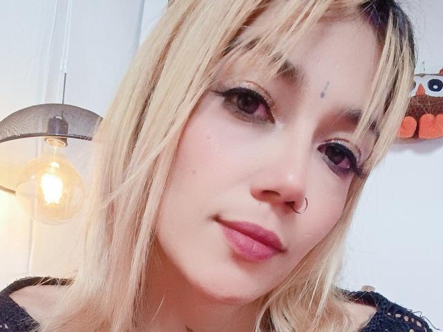 Veroo69, con cabello rubio, lápiz labial rosa, piercing en la nariz y delineador de ojos, sonriendo ligeramente. Tiene una pequeña marca en la piel sobre su ceja izquierda. En el fondo, hay una bombilla..