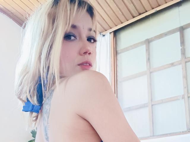 Veroo69 with blonde hair, blue hair tie, nose ring, and bare shoulders, looking over shoulder, slight smile, upper body, wooden ceiling, window in background.