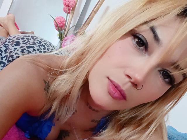 Veroo69, com cabelo loiro, lábios rosados e um piercing no nariz, vestindo uma blusa com estampa de leopardo, deitada em uma toalha rosa e azul, mostrando uma tatuagem no peito..