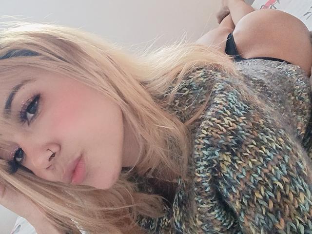 Veroo69 mit blonden Haaren, Nasenring und Eyeliner, liegt auf dem Bauch in einem grauen, gestrickten Pullover, wobei ihr Hintern in schwarzen Unterhosen zu sehen ist, und trägt einen schmollenden Gesichtsausdruck..